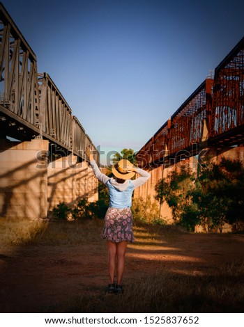 A slim  woman in a straw hat stands between two railways bridges hailing upcoming trains by her hand. Picture. 