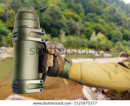 Photo of soldier hand in glove holding tactical thermos on nature background.