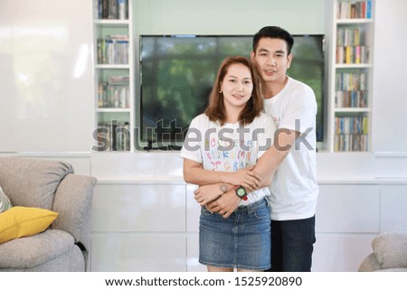 medium shot portrait of asian husband and asian wife hugging in living room and looking at camera with happy smiling face