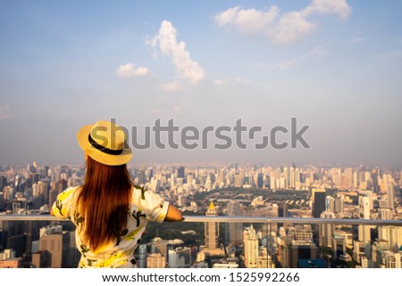 Woman wear yellow dress and hat, Asian traveler standing with their backs turned and looking aside on balcony with beautiful modern big city view on background.