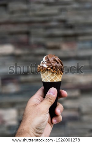Man holding vanilla and chocolate ice cream cone in hand, peanuts around cone, very blurred background, small depth of field. Dark background.
