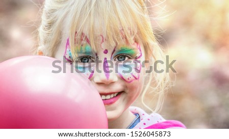 A girl dressed as a colourful butterfly holds a pink balloon and laughs happily into the camera