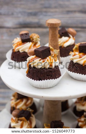 Chocolate cupcakes with cream cheese frosting, salted caramel sauce, brownie bites and toffees. Marble cakestand on grey wooden background.