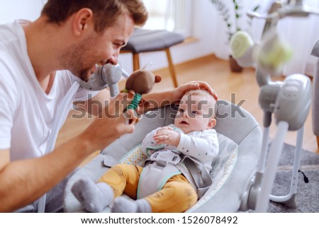 Happy adorable 6 months old baby boy lying in baby rocker chair and looking at toy that his caring dad holds. Healthy upbringing concept. Royalty-Free Stock Photo #1526078492