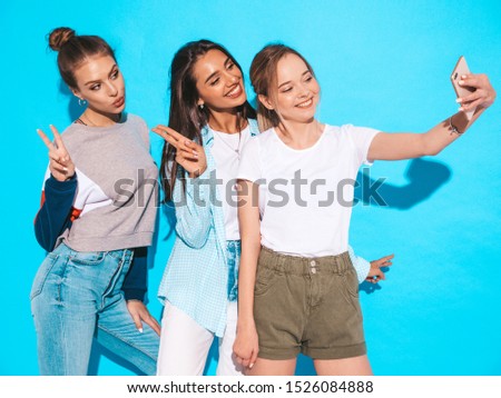 Three young smiling hipster women in summer clothes.Girls taking selfie self portrait photos on smartphone.Models posing near blue wall in studio,Female showing positive face emotions