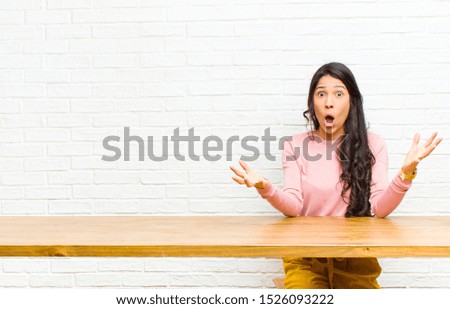 young  pretty latin woman open-mouthed and amazed, shocked and astonished with an unbelievable surprise sitting in front of a table