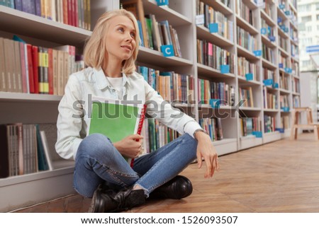 Attractive young woman smiling, looking away, sitting near bookshelves at the library, copy space. Charming happy female student sitting on the floor at college library