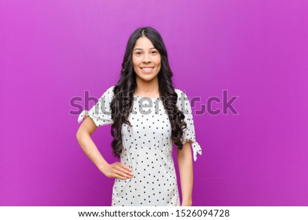 young pretty latin woman smiling happily with a hand on hip and confident, positive, proud and friendly attitude against purple wall