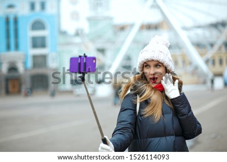 Emotional young woman wears warm coat and scarf taking selfie on her smart phone in winter during walk at the city