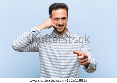 young handsome man smiling cheerfully and pointing to camera while making a call you later gesture, talking on phone