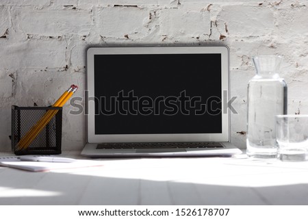Mock up empty black laptop screen on white brick wall background with pencils, notes and glass of water. Copyspace, negative space for your advertising, office and business style.