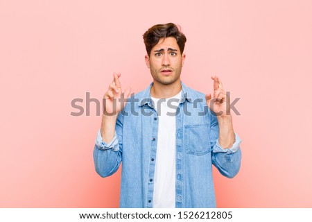 young handsome man crossing fingers anxiously and hoping for good luck with a worried look against pink background