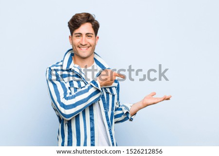 young handsome man smiling cheerfully and pointing to copy space on palm on the side, showing or advertising an object against blue background