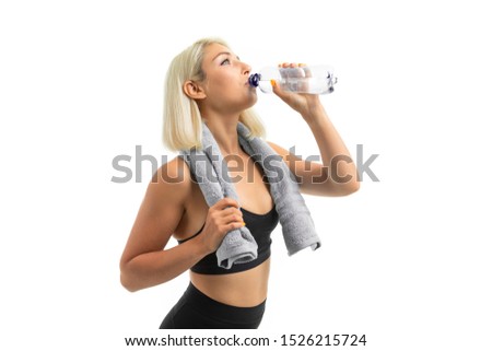 A young sports girl with blonde hair in a sports axe and black leggings holds a blue towel and a drinks a water.