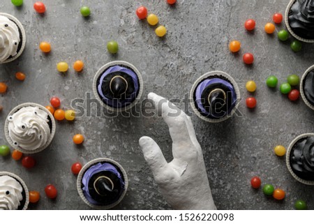 top view of decorative hand near delicious Halloween cupcakes with scattered colorful bonbons on concrete grey surface