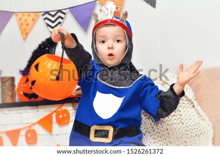 A little boy in a knight costume with a crown on his head sits with a pumpkin bucket for candies on a background of Halloween decorations.	