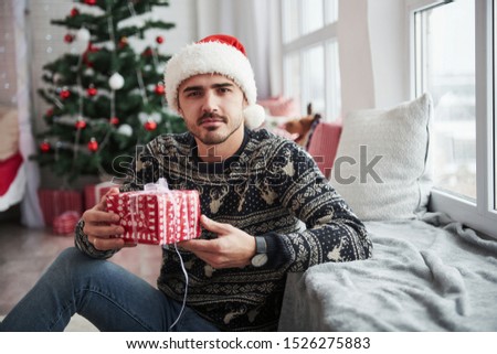 It's for you. Photo of man in santa hat and holiday clothes holds new year gift. Christmas tree on background.