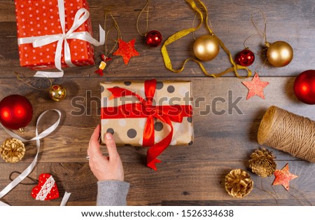 Person hands wrapping Christmas present on wooden background with toys, stars. Woman packaging wrapped gift box with red ribbon. New year holidays with gift boxes decoration, seasonal preparation.	