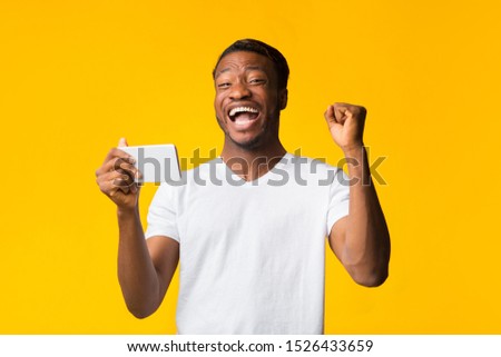 Excited Afro Man Holding Smartphone Shaking Fists Winning Mobile Game Standing Over Yellow Studio Background.
