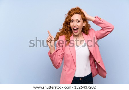 Redhead woman in suit over isolated blue wall surprised and pointing finger to the side