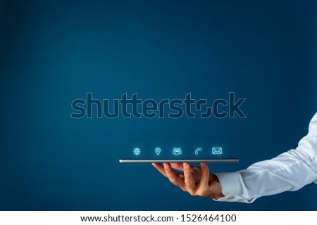 Male hand holding digital tablet with glowing interface contact and information icons coming out of it. Over navy blue background, with copy space.
