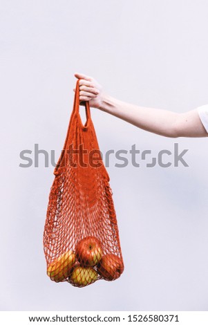 Female hand holds a red cotton shopping bag with oranges and apples on a background of white wall. The concept of naturalness and life with zero waste