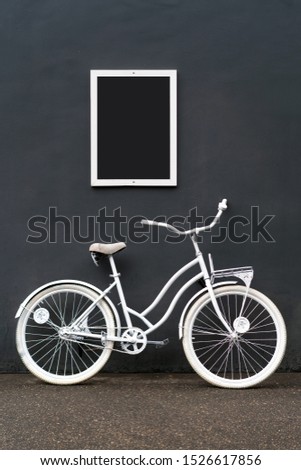 A white painted bicycle stands against a black wall. A white frame with a black empty space for inscriptions and pictures hangs on the wall. Minimalistic creative concept for creative ideas.