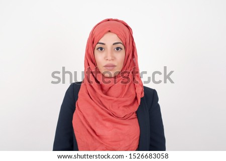 Waist up shot of joyful muslim girl looking to the camera, thinking about something. Both arms down, neutral facial expression. Standing outdoors wondering about something happening.
