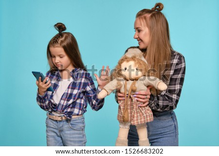 Mom and daughter dressed in checkered shirts and blue denim jeans are using smartphone while posing against a blue studio background. Close-up shot.