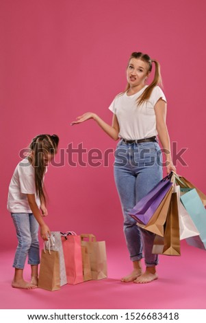 Mom and daughter with ponytails, dressed in white t-shirts and blue jeans are posing against pink background with packages in hands. Full length.