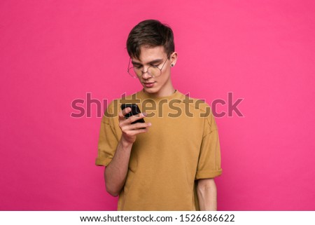 Guy in glasses and hat standing on a pink background. Looks at the phone screen
