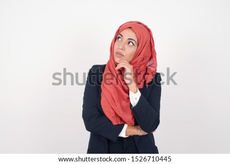 Portrait of thoughtful muslim girl keeps hand under chin, looks sideways, thinking or wondering about something with interest, dressed casually. Taking decisions concept.