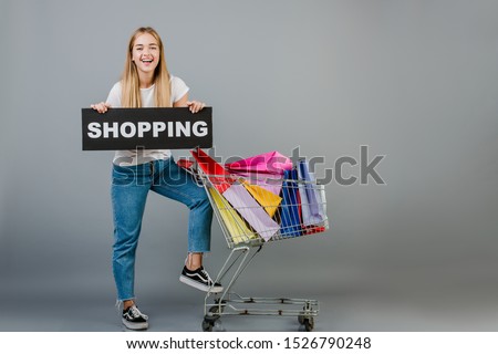 beautiful young blonde woman with shopping sign and pushcart with colorful shopping bags isolated over grey