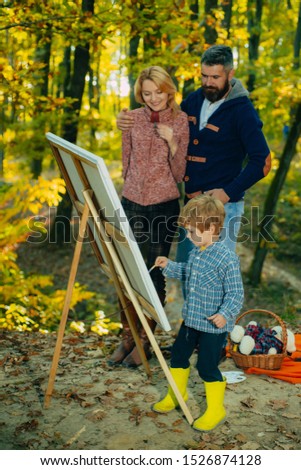 Blonde cute smart son drawing his family giving joyful moments to mom and dad. Parents selebreting anniversary. Family picnic. Mother, father, son drawing pictures together in green park. Family day