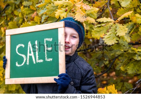 A boy in autumn clothes and a hat advertises the beginning of sales in stores. The boy is holding a sign that says discounts. Yellow leaves fall from trees in autumn.