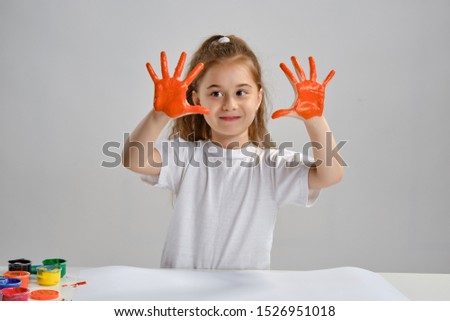 Little girl in white t-shirt sitting at table with whatman and colorful paints, showing her painted hands. Isolated on white. Medium close-up.