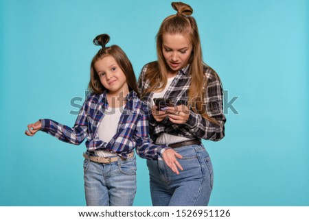 Mom and daughter dressed in checkered shirts and blue denim jeans are using smartphone while posing against a blue studio background. Close-up shot.