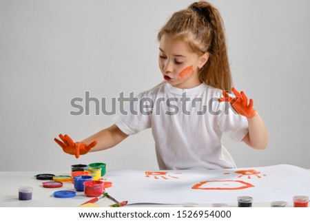 Little girl in white t-shirt sitting at table with whatman and colorful paints, painting on it with her hands. Isolated on white. Medium close-up.