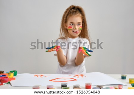 Little girl in white t-shirt sitting at table with whatman and colorful paints, showing her painted hands, face. Isolated on white. Medium close-up.