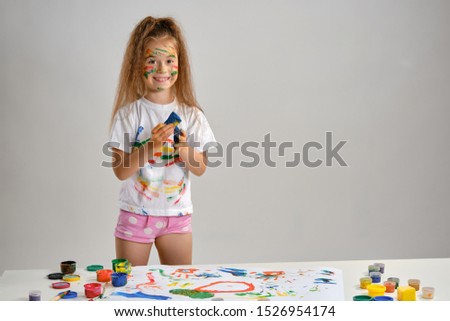 Little girl in white t-shirt standing at table with whatman and colorful paints, playing with a sponge soaked in paint. Isolated on white. Close-up.