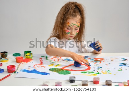 Little girl in white t-shirt sitting at table with whatman and paints, drawing on it, posing with painted face, hands. Isolated on white. Close-up.
