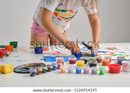 Little artist in white t-shirt standing at table with whatman and colorful paints, painting on it with her hands. Isolated on white. Close-up.