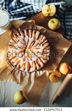 A round apple pie on parchment paper, lies on a wooden board among apples, next to a checkered towel and a glass of milk
