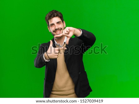 young businessman feeling happy, friendly and positive, smiling and making a portrait or photo frame with hands against green background