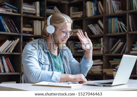Happy male student wear headphones waving hand looking at laptop computer screen video conference calling talking with online skype teacher communicating for distance education e learning in library Royalty-Free Stock Photo #1527046463
