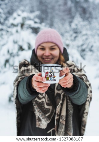 Young woman in pink hat demonstrating metal white cup with funny cartoon on snowy winter forest background. Hipster girl holding hot drink outdoors on nature. Active lifestyle leisure holidays