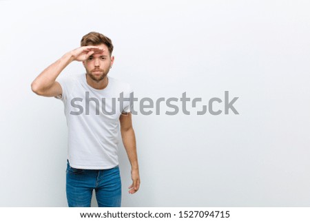 young man looking bewildered and astonished, with hand over forehead looking far away, watching or searching against white background