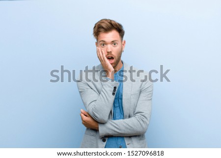 young businessman open-mouthed in shock and disbelief, with hand on cheek and arm crossed, feeling stupefied and amazed against blue background