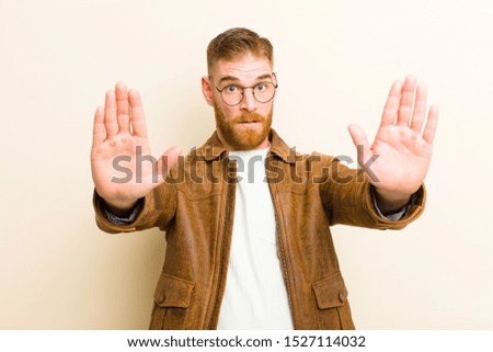 young red head man looking serious, unhappy, angry and displeased forbidding entry or saying stop with both open palms against beige background