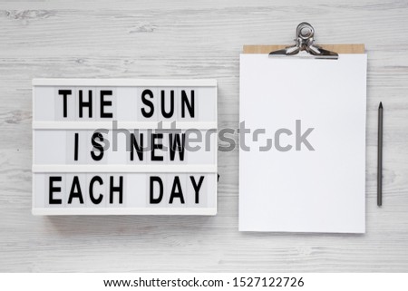 'The sun is new each day' words on a lightbox, clipboard with blank sheet of paper on a white wooden surface, top view. Overhead, from above, flat lay. Copy space.